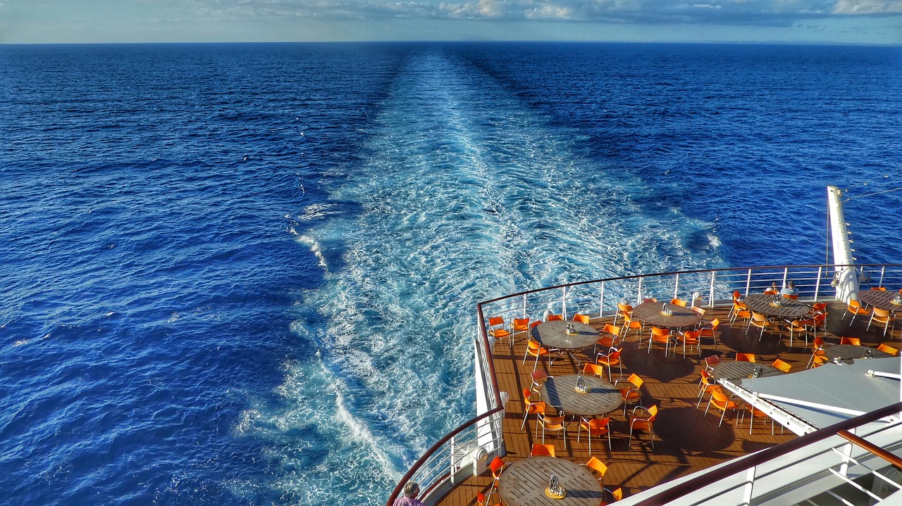 Stern view from the top deck of a luxury cruise ship, with all-inclusive dining tables set against the deep blue of the open ocean and the ships wake stretching into the horizon.