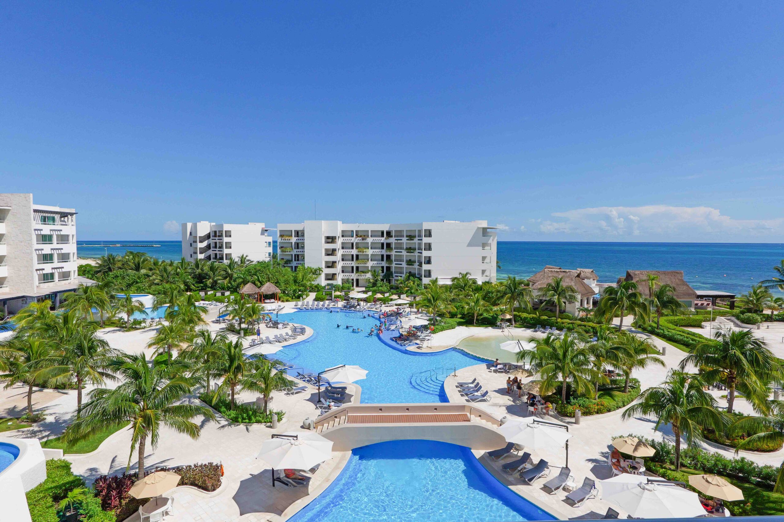 Balcony view of a luxury all-inclusive resort in Cancun, Mexico, with multilevel white hotels surrounding a massive pool, palm-filled gardens, with Caribbean Ocean side bungalows serving all-inclusive drinks.