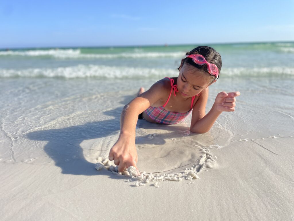 Family travel moment in Destin, Florida — daughter drawing in the white sand while parent works from home on the beach.