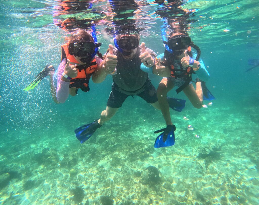 Family snorkeling at the Underwater Museum in Isla Mujeres, Mexico — father and kids floating together with thumbs up in crystal-clear Caribbean water.
