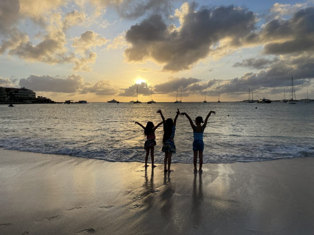Three daughters lifting their hands towards the sky in praise to Jesus Christ at sunset on Simpson Bay, Sint Maarten, as the Caribbean Ocean stretches to the horizon with sailboats dotting the distant horizon.