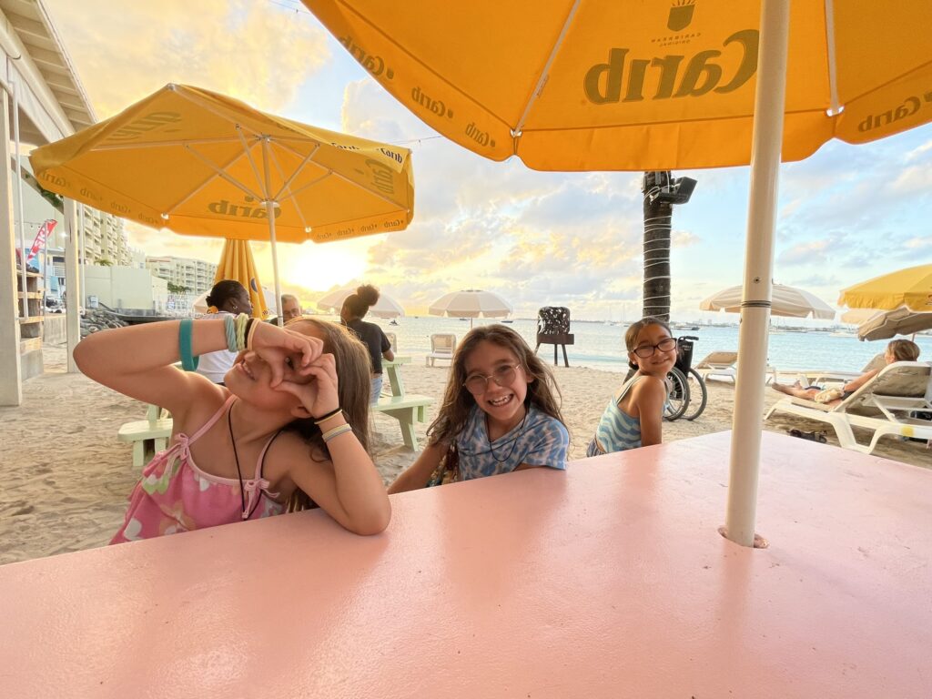 Family sunset at Simpson Bay, St. Maarten — three children at a picnic table by the beach, smiling with ocean views and umbrellas along the horizon.
