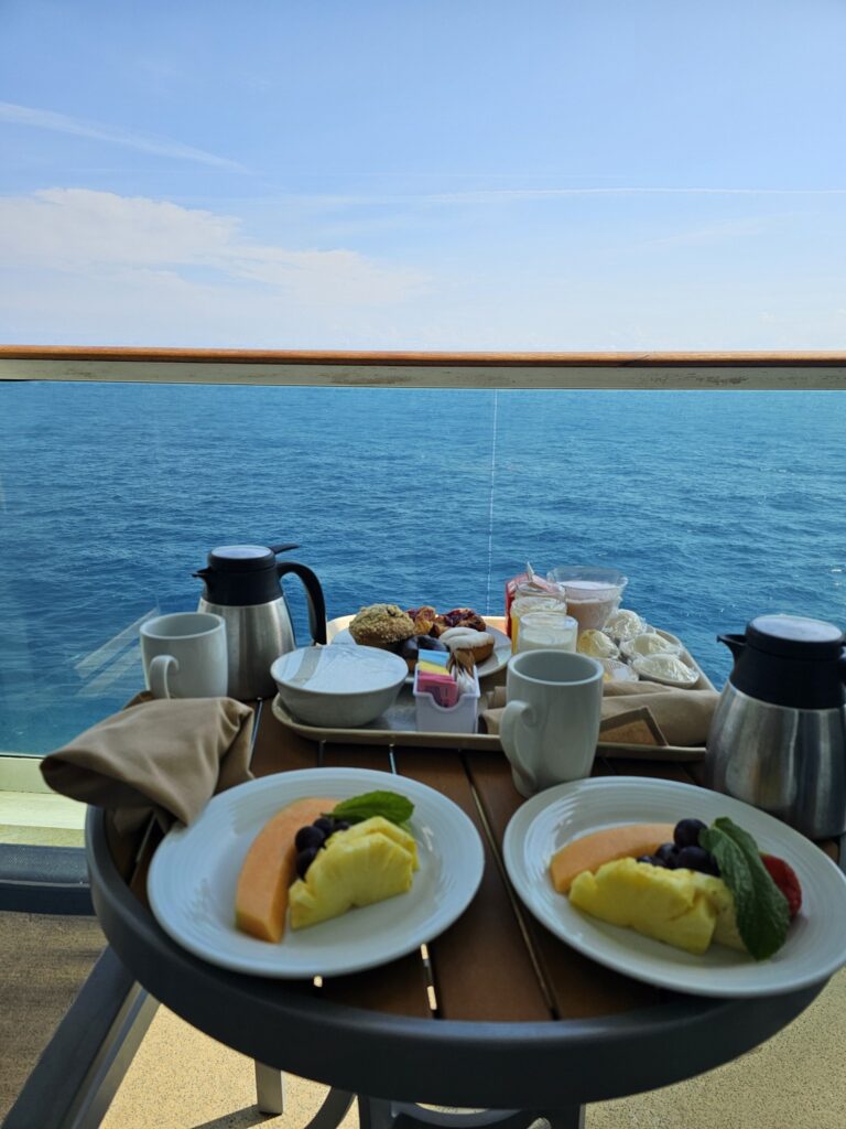 View of ocean on cruise ship balcony where breakfast is sitting on a small table against the glass railing with the ocean extending into the horizon representing a luxury cruise into your dream vacation ocean view balcony experience.