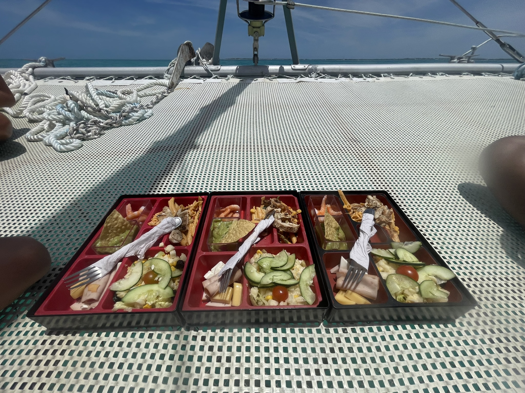 Lunch platter served aboard a Cancún Adventures catamaran, featuring shrimp cocktail, chicken with noodles, chips and guacamole, ham and cheese, and a fresh salad on the front bridge netting.