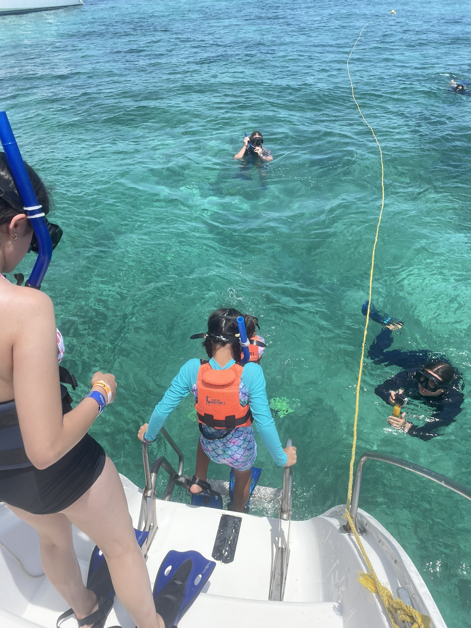 Child wearing a life jacket and snorkel gear stepping down the swim platform stairs, with dad waiting nearby and a yellow tow line extending into the water to keep the group safely positioned before snorkeling.