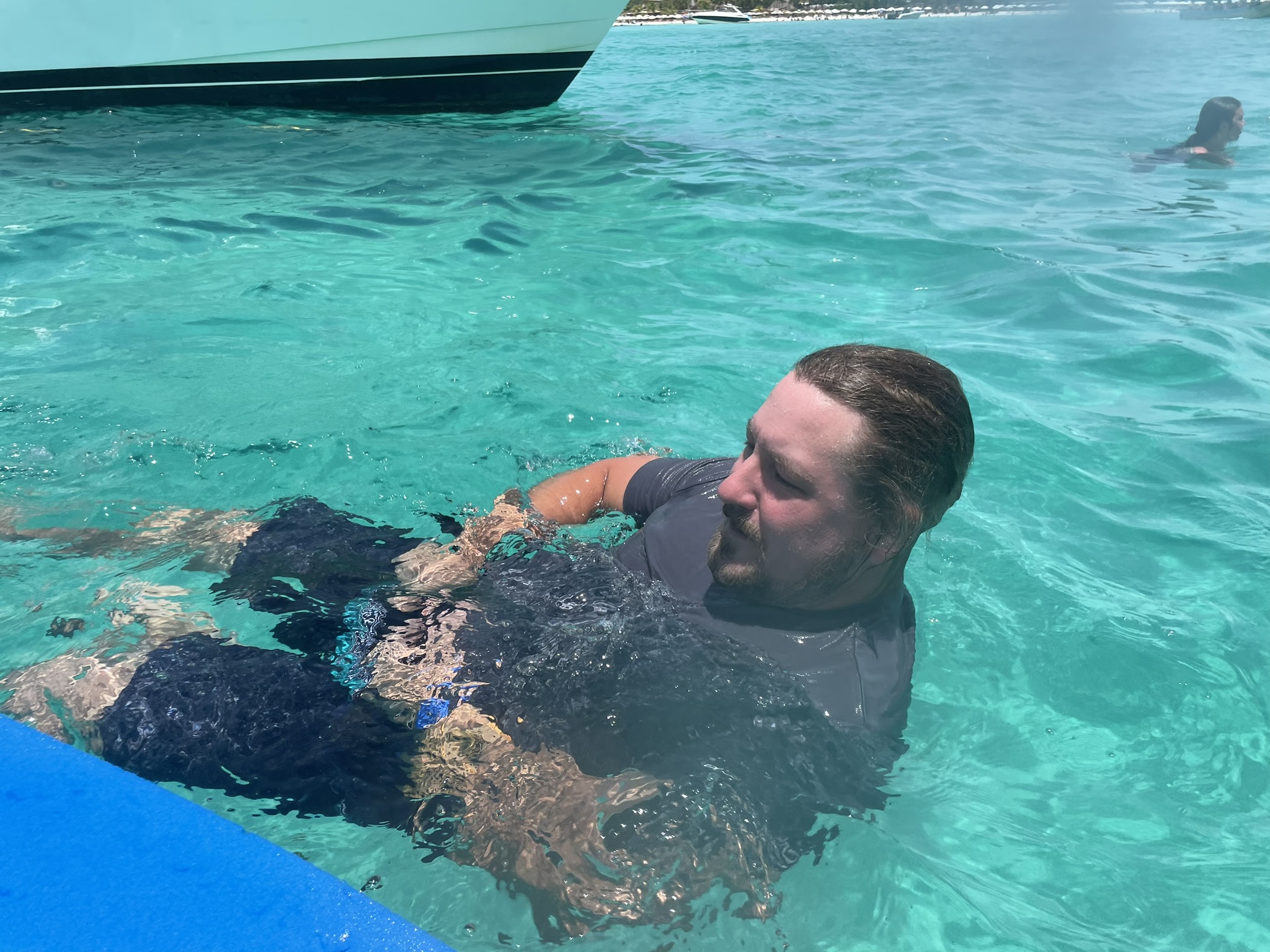 Man floating beside a swim pad in 40-foot clear Caribbean water near Isla Mujeres reef, surrounded by other boats of tourists during the after-snorkeling cooldown.