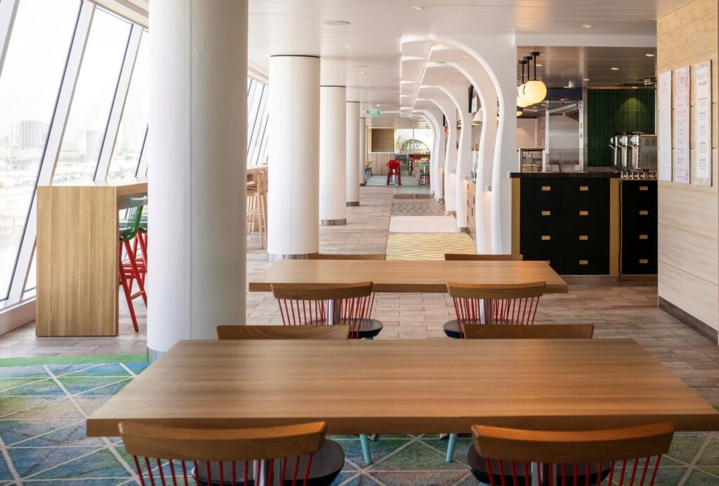 Hall view of the Icon ship class Aqua Dome Market Side eatery table area with window views and buffet style chiefs' area representing endless all you can eat dining experiences while you cruise into your dream vacation.