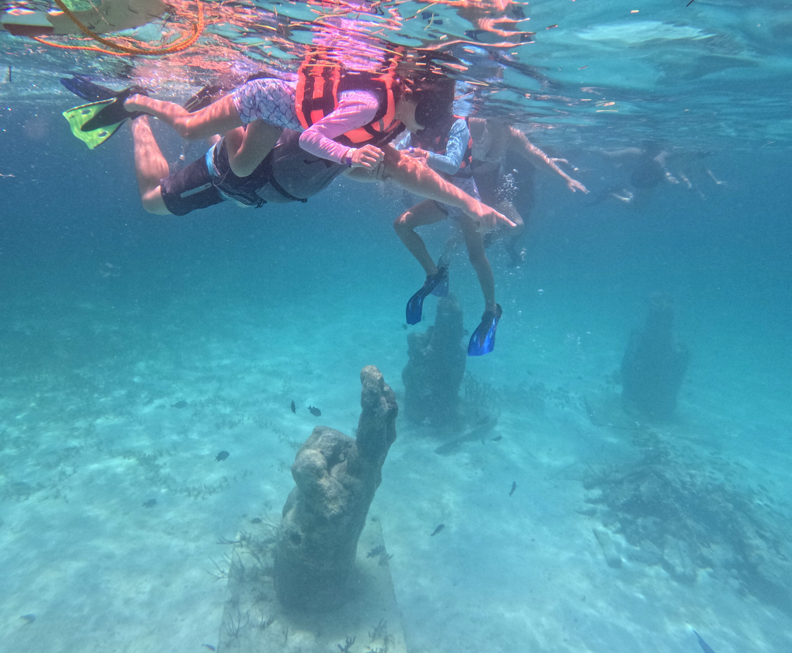 Parent and two children snorkeling above underwater statues at the MUSA Ocean Art Museum near Isla Mujeres, Mexico, surrounded by 30-foot-deep crystal-clear Caribbean water.