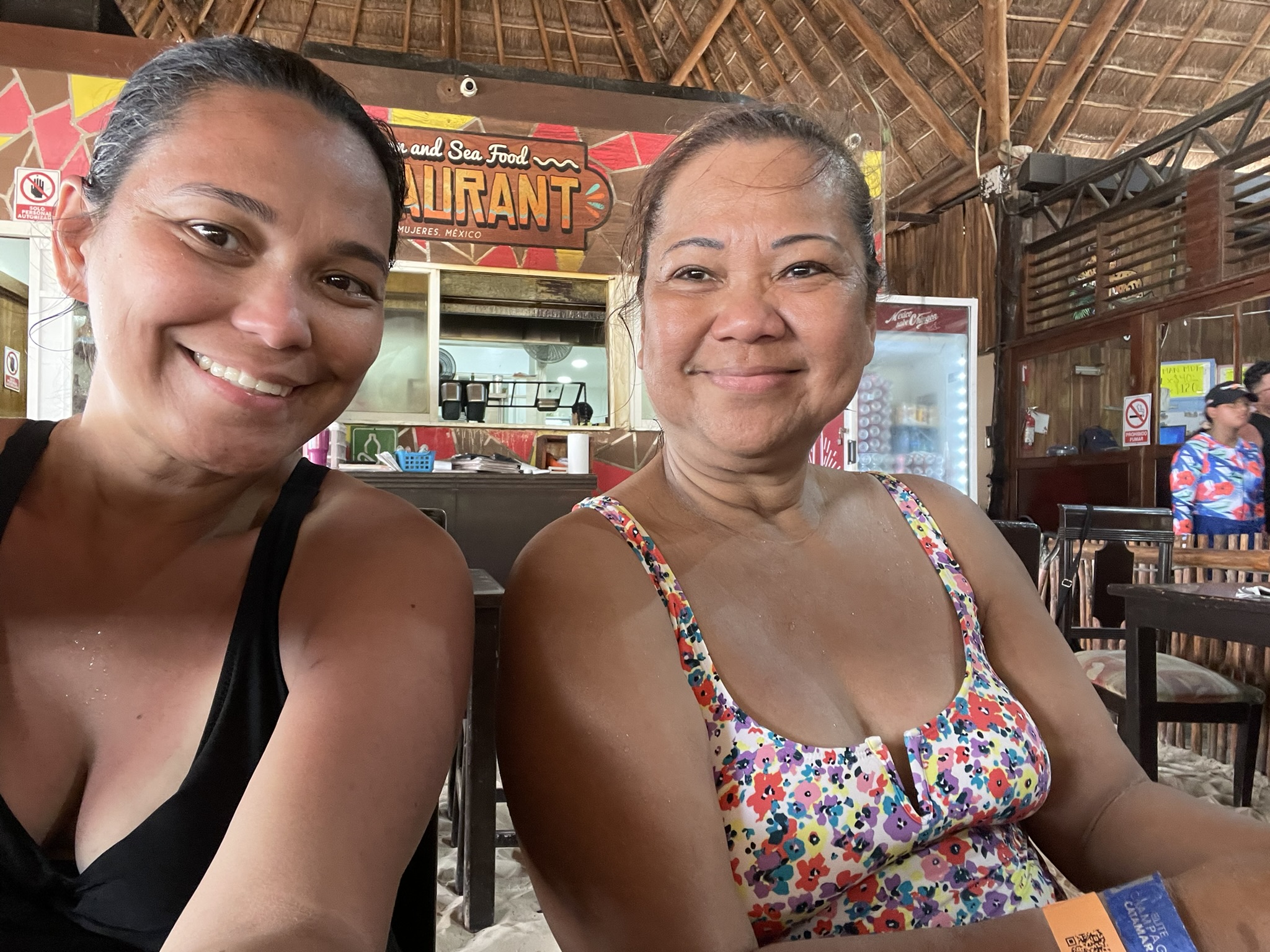 Mother and daughter sitting together at a table in the Perl Negra (Tzalam Mexican Food & Sea) restaurant, smiling and sunburned after a day of sailing and snorkeling, with the restaurant’s sign visible in the background.