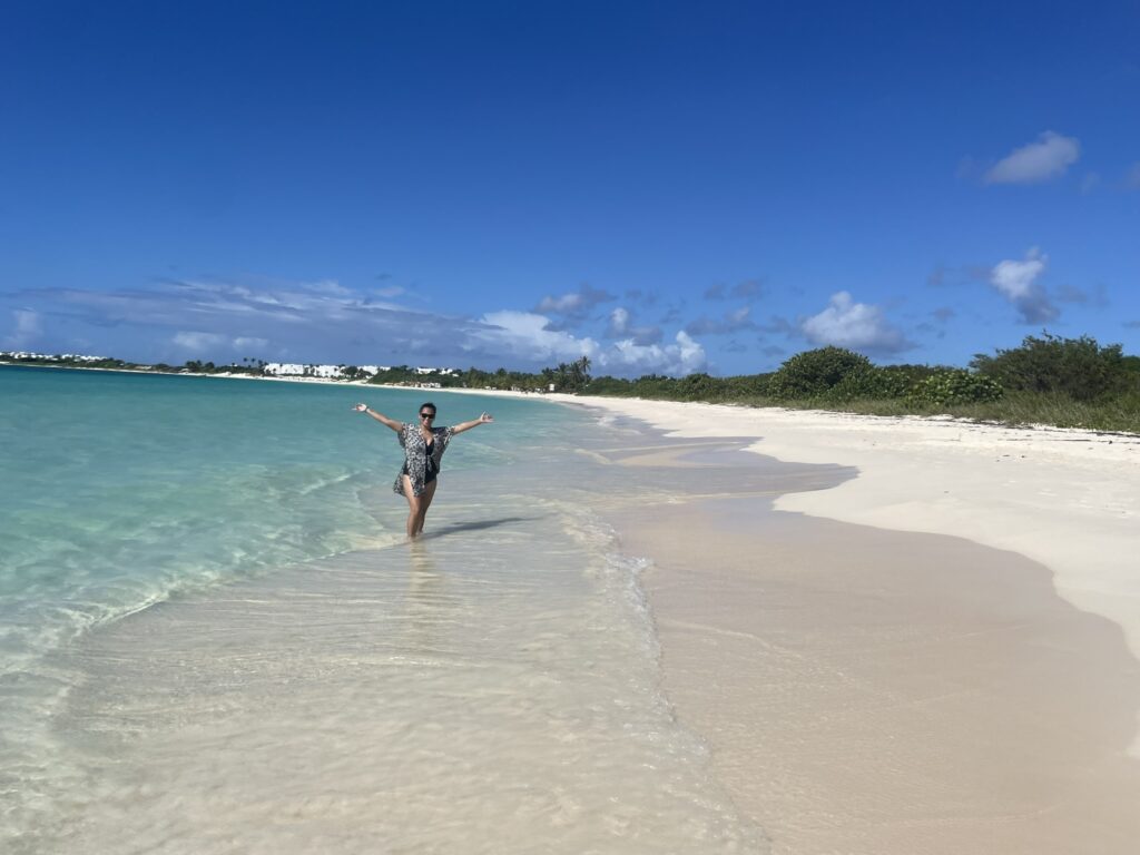Traveler standing with arms raised on the white sand shoreline of Rendezvous Bay Anguilla with curved beach stretching into the distance.