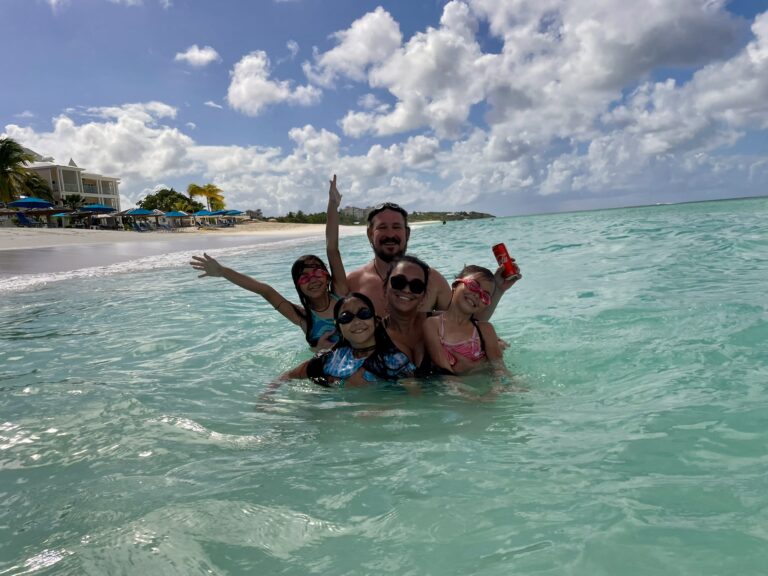 Family swimming together in the crystal-clear water at Shoal Bay Anguilla with beach umbrellas and resorts visible along the shoreline.
